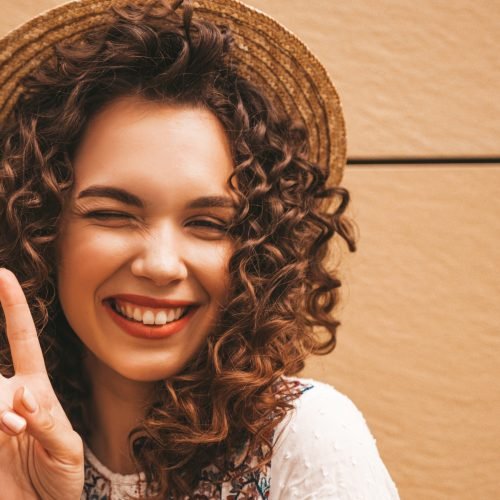 Beautiful smiling model with afro curls hairstyle dressed in summer hipster white dress.Sexy carefree girl posing in the street near yellow wall in hat.Funny and positive woman having fun