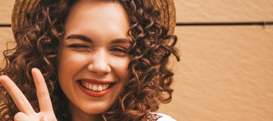 Beautiful smiling model with afro curls hairstyle dressed in summer hipster white dress.Sexy carefree girl posing in the street near yellow wall in hat.Funny and positive woman having fun