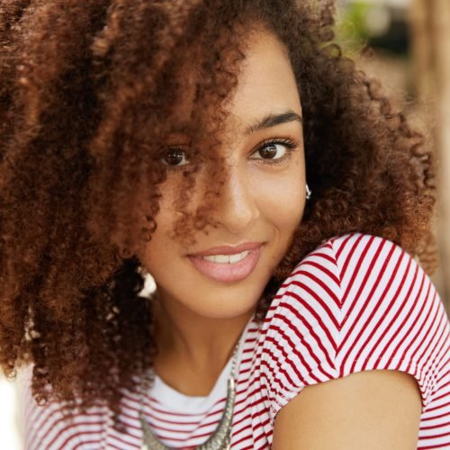 Close up shot of lovely dark skinned mixed race female with wavy dark hair, smiles pleasantly, wears striped t shirt, being satisfied with everything. Adorable African woman expresses happiness